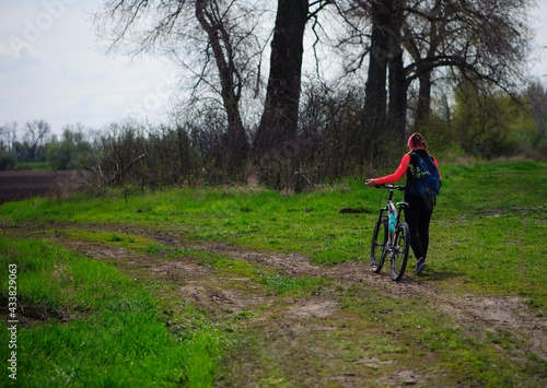 a girl with a bicycle walks a field trail. Woman riding a mountain bike in the forest. Hiker woman with backpack. one young woman riding a bike in nature. rolling the bike in his hands. back view.