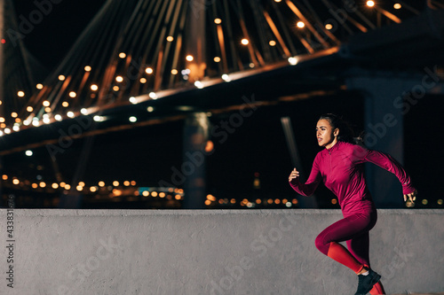 Woman running at night on embankment. Side view of female jogger sprinting outdoors against bridge. © Artem Varnitsin