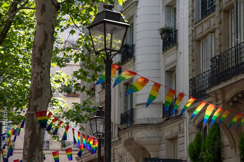 Decoration of triangle shape banners in colors of Lgbtq flags hanging between vintage lantern streetlights and ornate stone house with balconies. Gay pride parade in Paris France