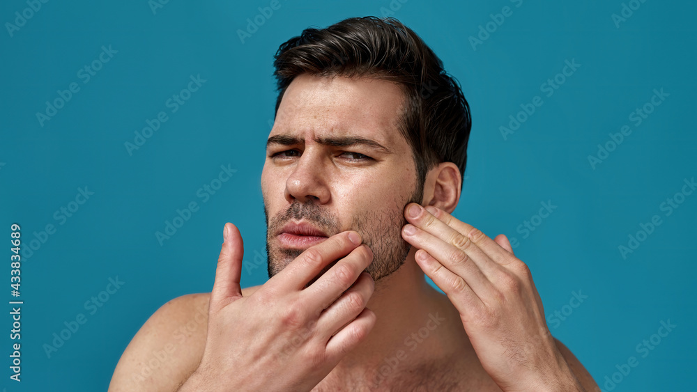 Fototapeta premium Portrait of shirtless handsome brunette guy checking his skin, looking anxious, posing isolated over blue background