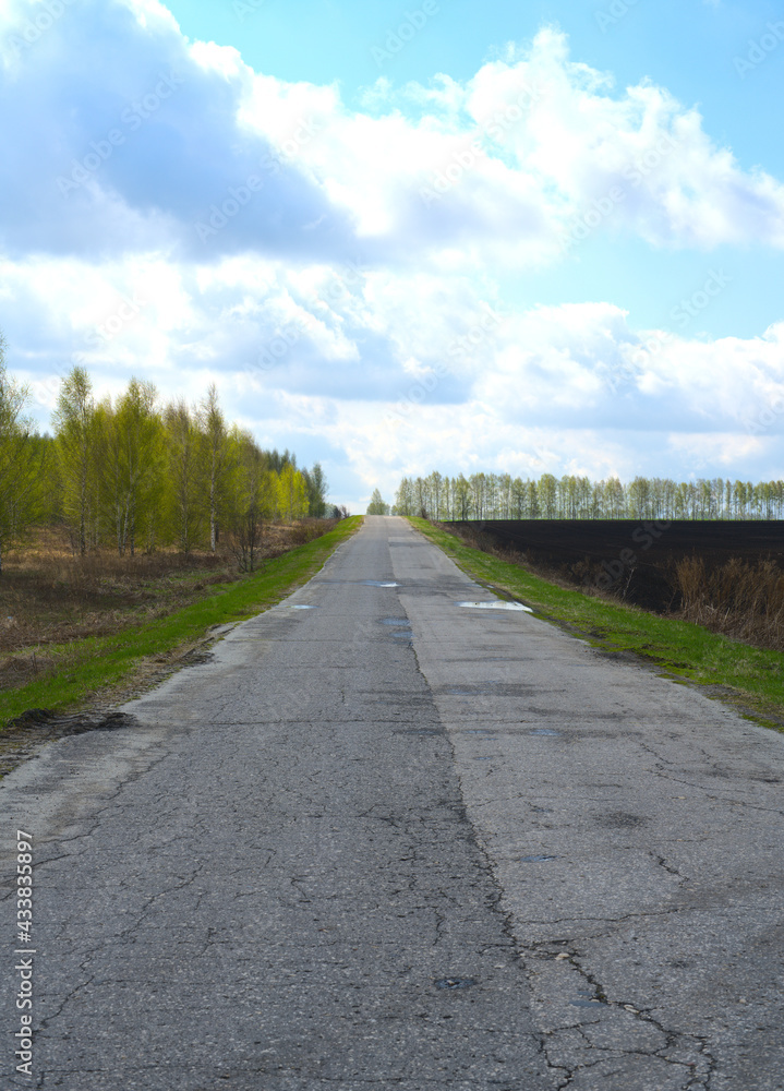 A broken road stretching away into the distance. To the left is a forest and to the right is a plowed agricultural field. Spring A broken road stretching away into the distance. To the left is a forest and to the right is a plowed agricultural field. Spring