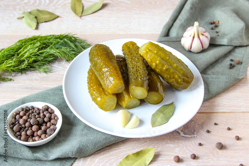 Plate of pickled homemade cucumbers on wooden rustic background. Marinated pickled cucumbers on the white plate with garlic, black pepper and bay leaf. Food concept. Homemade pickled vegetables.