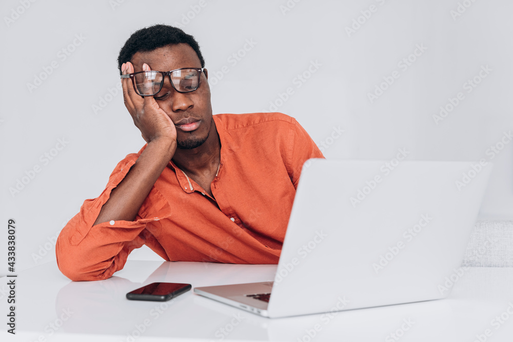 African man in an orange shirt and glasses sleeps leaning on his arm sitting at his desk in front of a laptop and phone on a light background. Young freelancer tired of working on a project, a break African man in an orange shirt and glasses sleeps leaning on his arm sitting at his desk in front of a laptop and phone on a light background. Young freelancer tired of working on a project, a break