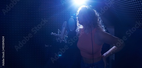 Beautiful blonde girl emotionally singing song in recording studio with professional microphone and headphones  creates new track album  vocal artist silhouette in blue light
