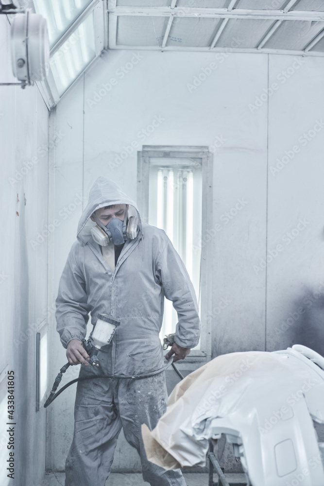 Car painting and automobile repair service. Auto mechanic in white overalls paints car with airbrush pulverizer in paint chamber.