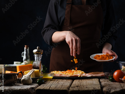 Steak with vegetables and cheese. Cooking process. The cook sprinkles carrots on the pieces of meat. Rough wooden table. The ingredients on the table are cheese, garlic, lemon, salt. Dark tones.