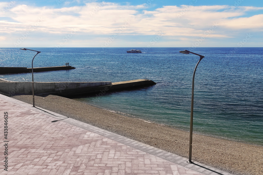 Sea Paved Tiled Promenade Or Pedestrian Path With Led Lamppost And Pebble Beach With Breakwaters Pier With Two Adult Couple Who Look At The Boat And Fishing In The Sea Water. 