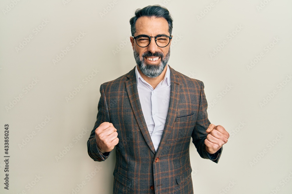 Middle age man with beard and grey hair wearing business jacket and glasses excited for success with arms raised and eyes closed celebrating victory smiling. winner concept. Middle age man with beard and grey hair wearing business jacket and glasses excited for success with arms raised and eyes closed celebrating victory smiling. winner concept.