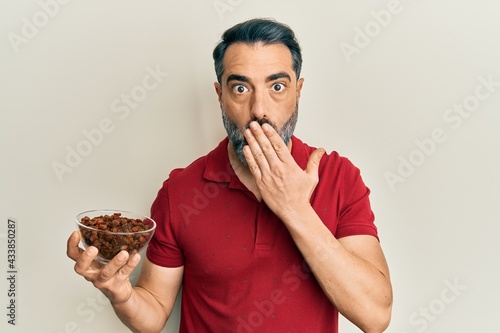 Middle age man with beard and grey hair holding raisins bowl covering mouth with hand  shocked and afraid for mistake. surprised expression