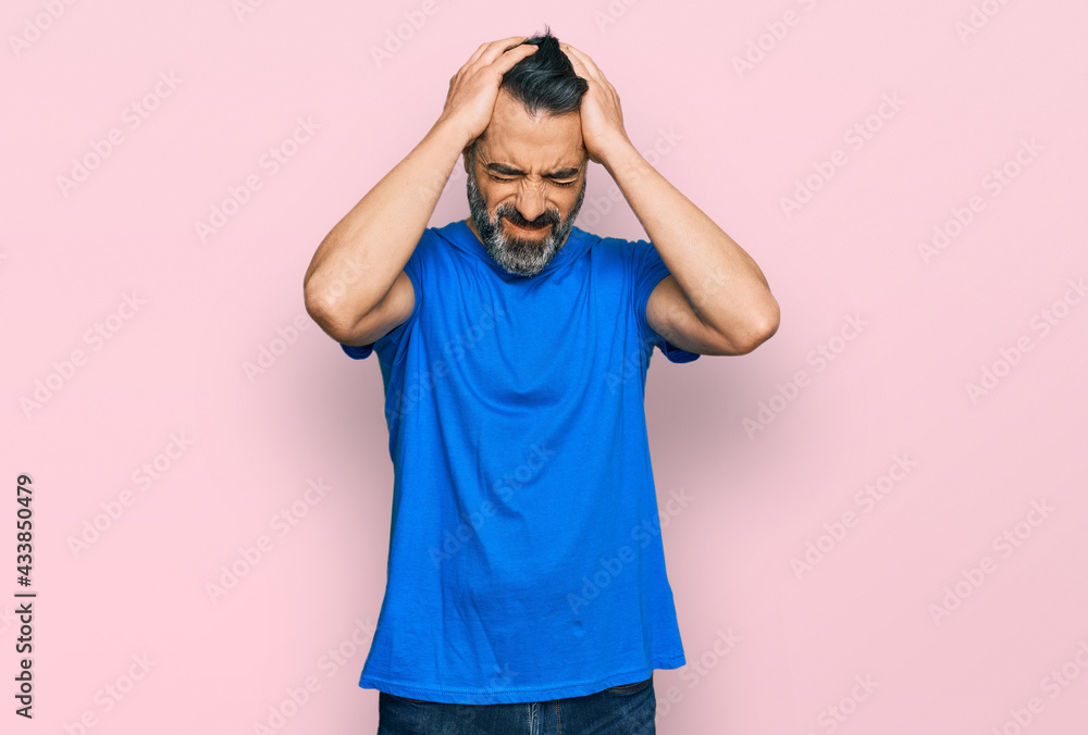 Middle aged man with beard wearing casual blue t shirt suffering from headache desperate and stressed because pain and migraine. hands on head. Middle aged man with beard wearing casual blue t shirt suffering from headache desperate and stressed because pain and migraine. hands on head.