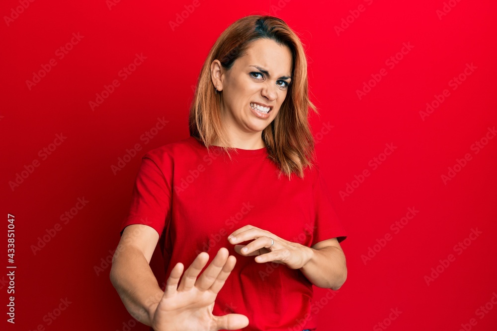 Naklejka premium Hispanic young woman wearing casual red t shirt disgusted expression, displeased and fearful doing disgust face because aversion reaction. with hands raised