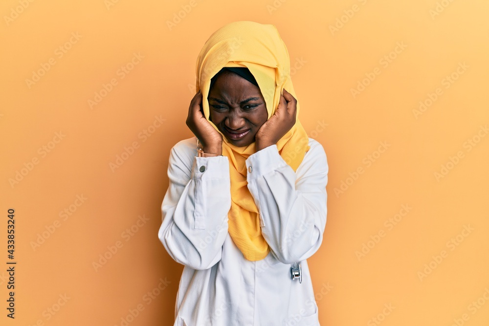 Beautiful african young woman wearing doctor uniform and hijab covering ears with fingers with annoyed expression for the noise of loud music. deaf concept. Beautiful african young woman wearing doctor uniform and hijab covering ears with fingers with annoyed expression for the noise of loud music. deaf concept.