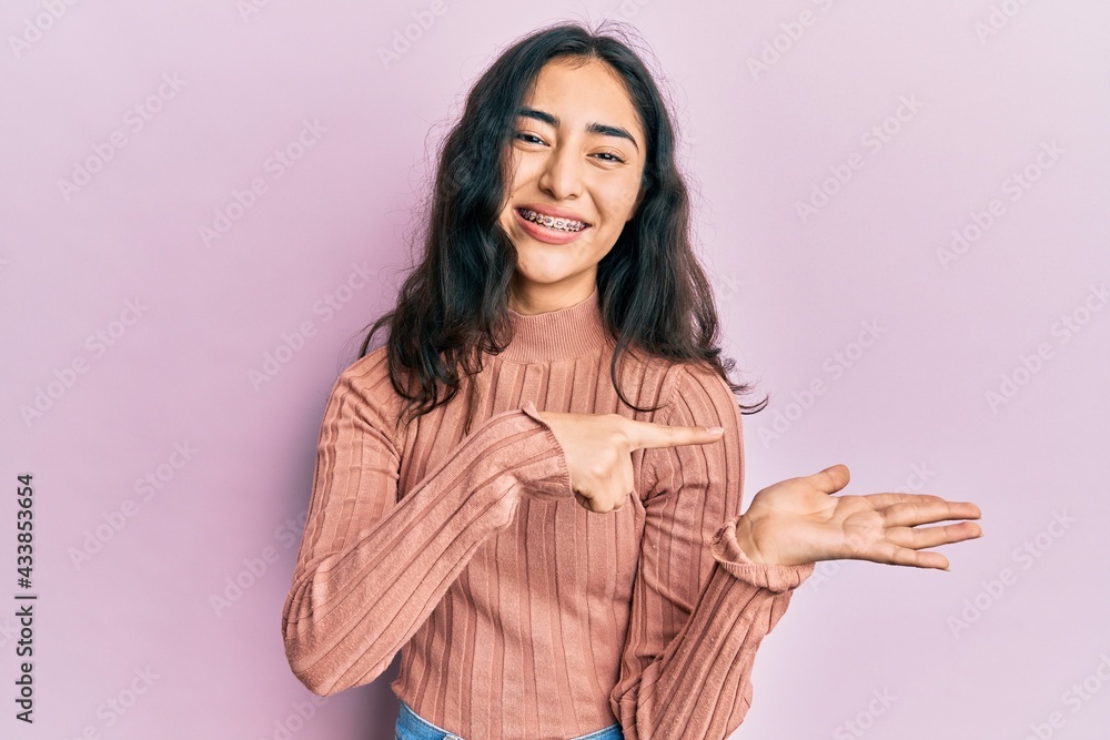 Hispanic teenager girl with dental braces wearing casual clothes amazed and smiling to the camera while presenting with hand and pointing with finger. Hispanic teenager girl with dental braces wearing casual clothes amazed and smiling to the camera while presenting with hand and pointing with finger.