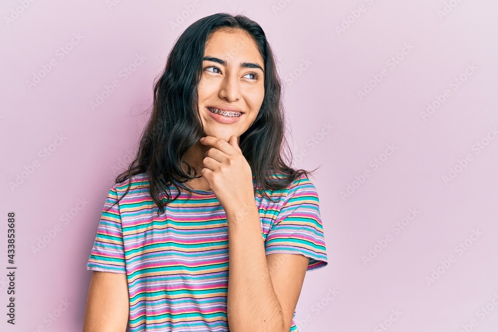 Hispanic teenager girl with dental braces wearing casual clothes with hand on chin thinking about question, pensive expression. smiling with thoughtful face. doubt concept. Hispanic teenager girl with dental braces wearing casual clothes with hand on chin thinking about question, pensive expression. smiling with thoughtful face. doubt concept.