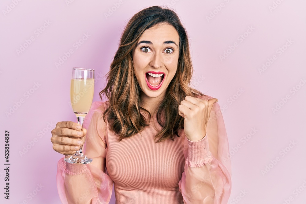 Young hispanic girl drinking a glass of sparkling champagne pointing thumb up to the side smiling happy with open mouth Young hispanic girl drinking a glass of sparkling champagne pointing thumb up to the side smiling happy with open mouth