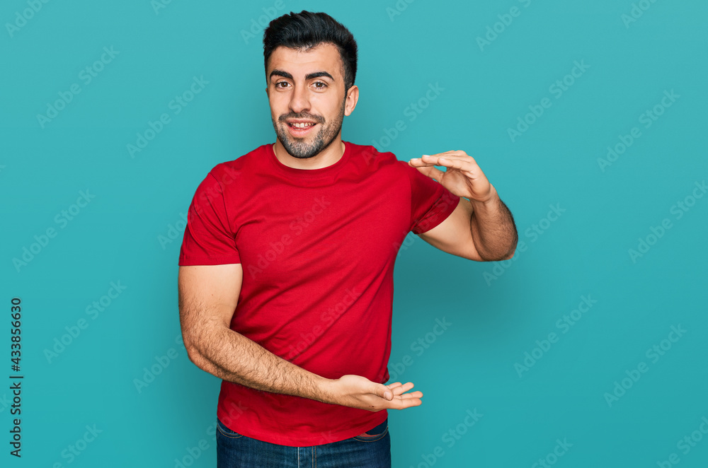 Fototapeta premium Hispanic man with beard wearing casual red t shirt gesturing with hands showing big and large size sign, measure symbol. smiling looking at the camera. measuring concept.