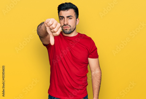 Hispanic man with beard wearing casual red t shirt looking unhappy and angry showing rejection and negative with thumbs down gesture. bad expression.