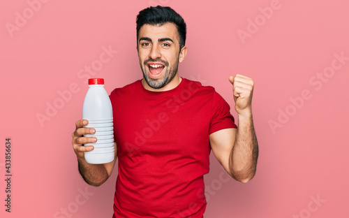 Hispanic man with beard holding liter bottle of milk screaming proud, celebrating victory and success very excited with raised arms