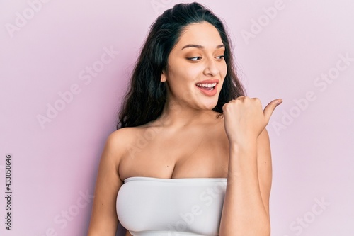 Beautiful middle eastern woman standing wearing a top showing skin pointing thumb up to the side smiling happy with open mouth