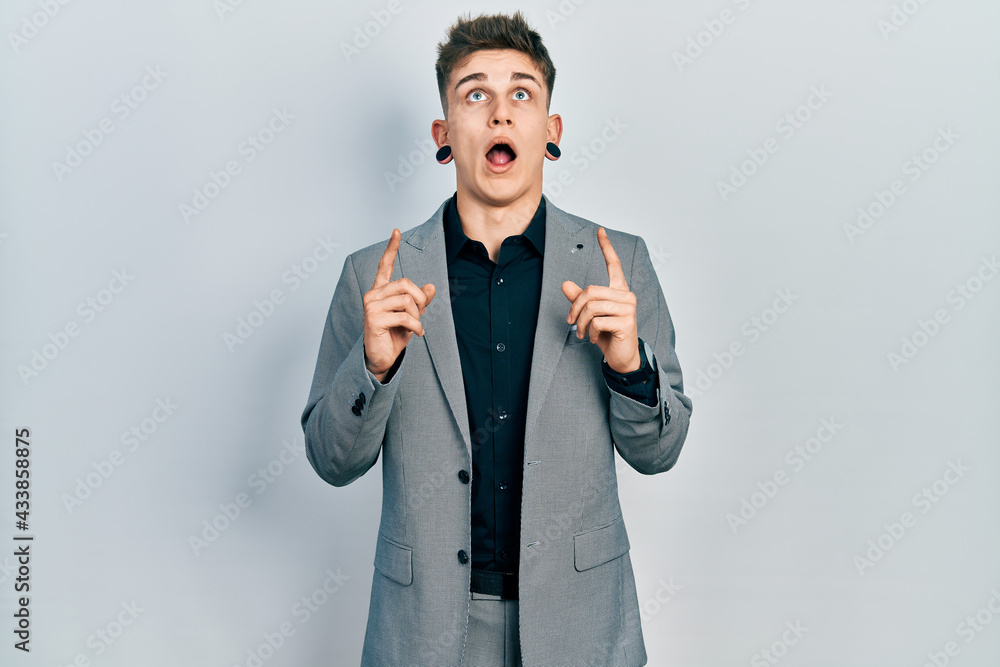 Young caucasian boy with ears dilation wearing business jacket amazed and surprised looking up and pointing with fingers and raised arms. Young caucasian boy with ears dilation wearing business jacket amazed and surprised looking up and pointing with fingers and raised arms.