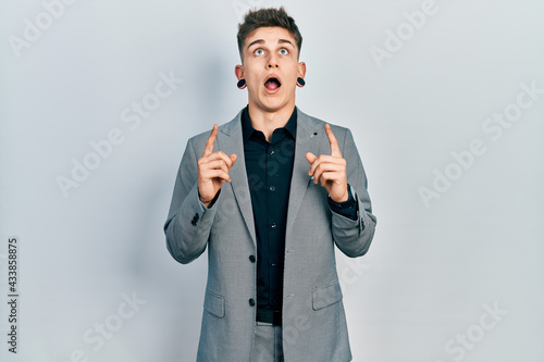 Young caucasian boy with ears dilation wearing business jacket amazed and surprised looking up and pointing with fingers and raised arms.