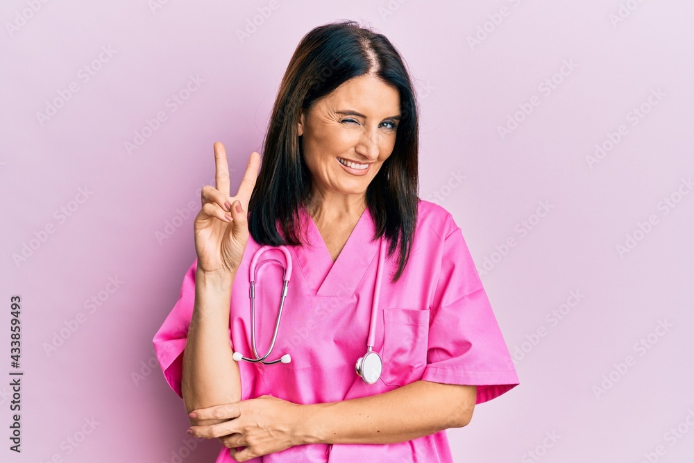 Middle age brunette woman wearing doctor uniform and stethoscope smiling with happy face winking at the camera doing victory sign. number two. Middle age brunette woman wearing doctor uniform and stethoscope smiling with happy face winking at the camera doing victory sign. number two.