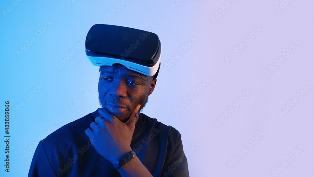 African-American guy with virtual reality glasses on his forehead looking around. Thinking something. Virtual headset glasses. Blue background. Studio footage. . High quality photo African-American guy with virtual reality glasses on his forehead looking around. Thinking something. Virtual headset glasses. Blue background. Studio footage. . High quality photo