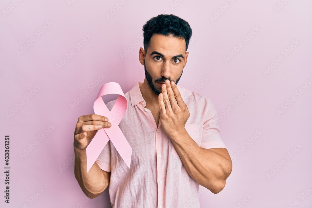 Young hispanic man holding pink cancer ribbon covering mouth with hand, shocked and afraid for mistake. surprised expression Young hispanic man holding pink cancer ribbon covering mouth with hand, shocked and afraid for mistake. surprised expression