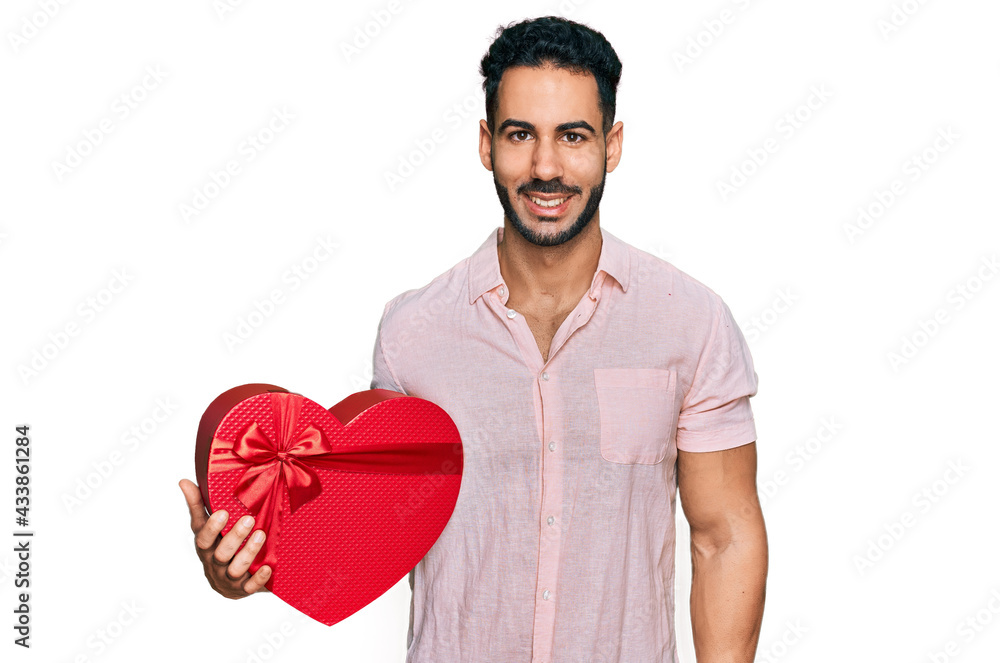 Hispanic man with beard holding valentine gift looking positive and happy standing and smiling with a confident smile showing teeth Hispanic man with beard holding valentine gift looking positive and happy standing and smiling with a confident smile showing teeth