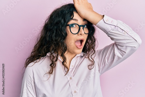 Young brunette woman with curly hair wearing casual clothes and glasses surprised with hand on head for mistake, remember error. forgot, bad memory concept.