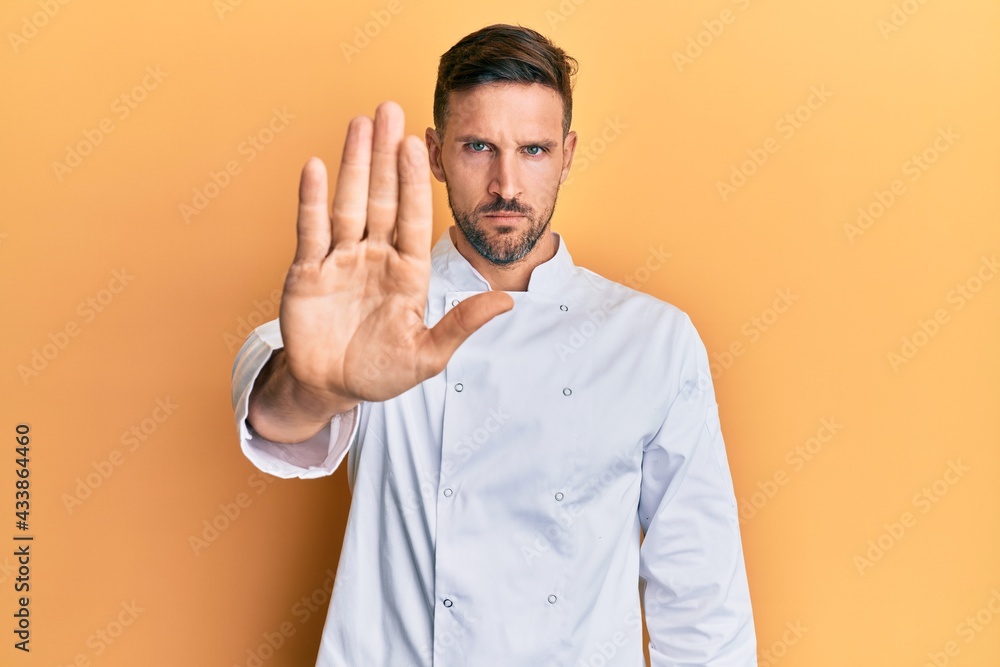 Handsome man with beard wearing professional cook uniform doing stop sing with palm of the hand. warning expression with negative and serious gesture on the face. Handsome man with beard wearing professional cook uniform doing stop sing with palm of the hand. warning expression with negative and serious gesture on the face.