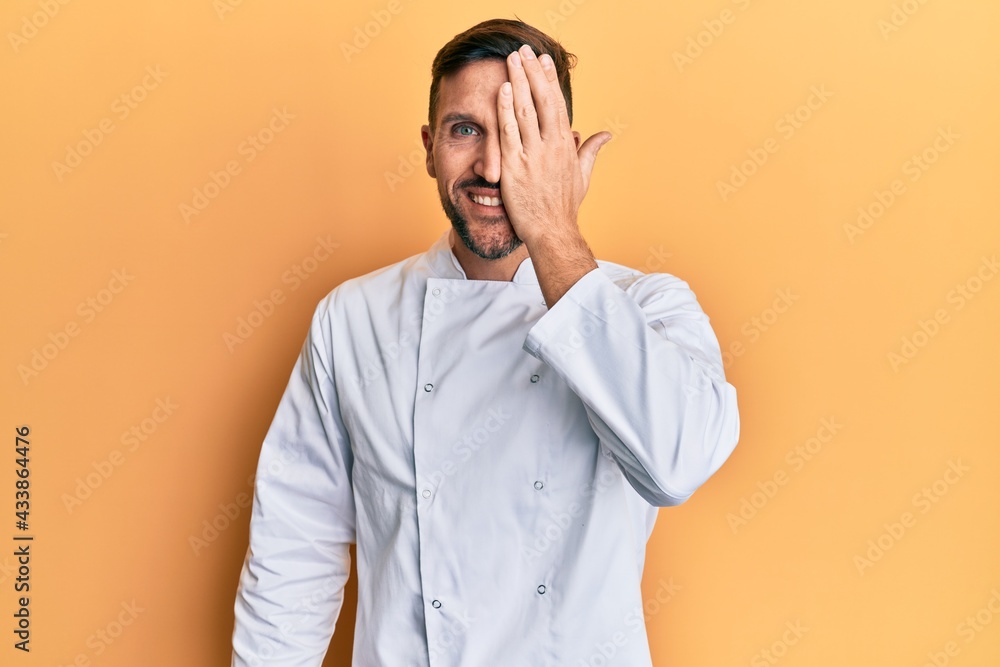 Handsome man with beard wearing professional cook uniform covering one eye with hand, confident smile on face and surprise emotion. Handsome man with beard wearing professional cook uniform covering one eye with hand, confident smile on face and surprise emotion.
