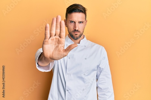 Handsome man with beard wearing professional cook uniform doing stop sing with palm of the hand. warning expression with negative and serious gesture on the face. © Krakenimages.com