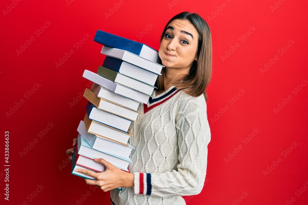 Young brunette student girl holding a pile of books puffing cheeks with funny face. mouth inflated with air, catching air. Young brunette student girl holding a pile of books puffing cheeks with funny face. mouth inflated with air, catching air.