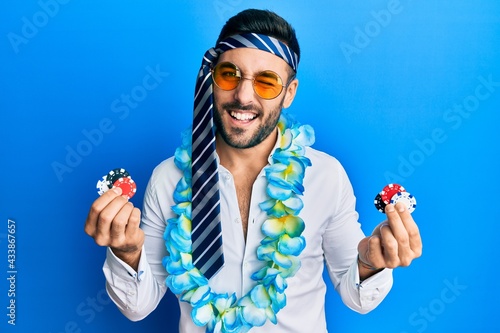 Young hispanic businessman wearing party funny style holding casino chips winking looking at the camera with sexy expression, cheerful and happy face. photo