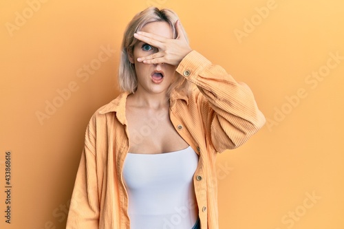 Young blonde girl wearing casual clothes peeking in shock covering face and eyes with hand, looking through fingers with embarrassed expression.