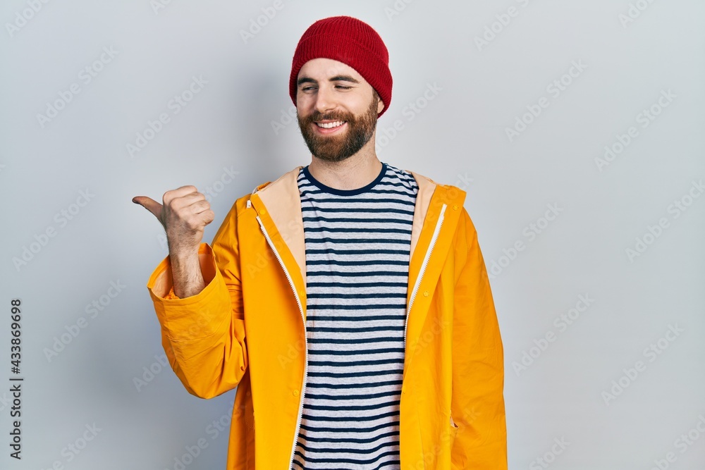 Caucasian man with beard wearing yellow raincoat smiling with happy face looking and pointing to the side with thumb up. Caucasian man with beard wearing yellow raincoat smiling with happy face looking and pointing to the side with thumb up.