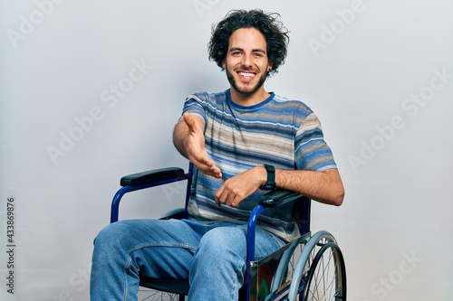 Handsome hispanic man sitting on wheelchair smiling friendly offering handshake as greeting and welcoming. successful business.