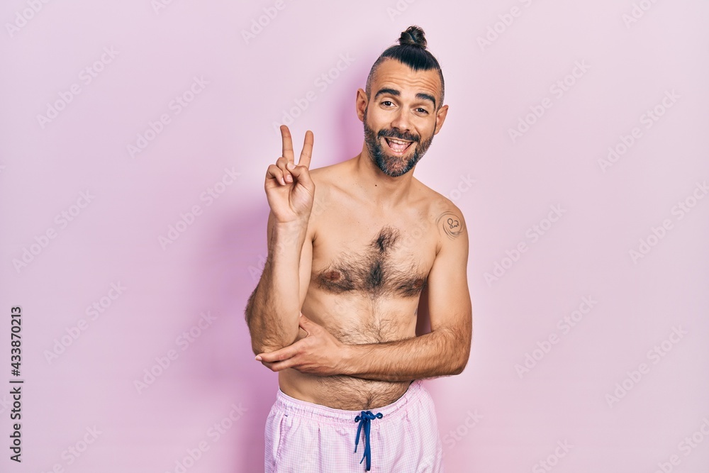 Young hispanic man shirtless wearing swimsuit smiling with happy face winking at the camera doing victory sign. number two.