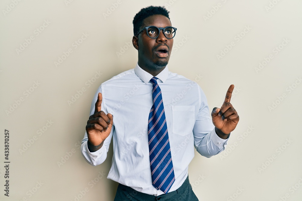 Handsome black man wearing glasses business shirt and tie amazed and surprised looking up and pointing with fingers and raised arms. Handsome black man wearing glasses business shirt and tie amazed and surprised looking up and pointing with fingers and raised arms.