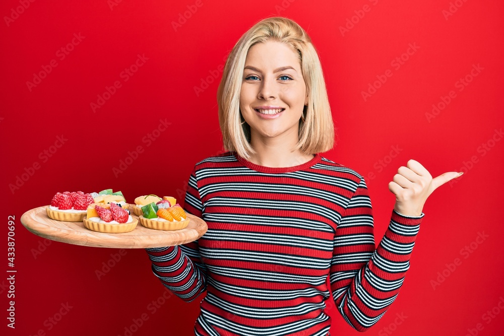 Young blonde woman holding sweet pastries pointing thumb up to the side smiling happy with open mouth