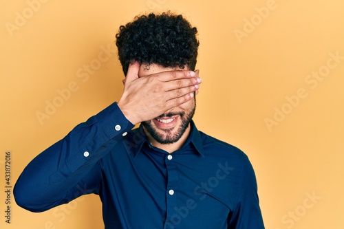 Young arab man with beard wearing casual shirt smiling and laughing with hand on face covering eyes for surprise. blind concept.