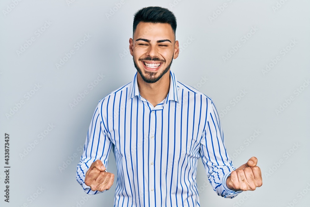 Young hispanic man with beard wearing casual striped shirt excited for success with arms raised and eyes closed celebrating victory smiling. winner concept. Young hispanic man with beard wearing casual striped shirt excited for success with arms raised and eyes closed celebrating victory smiling. winner concept.