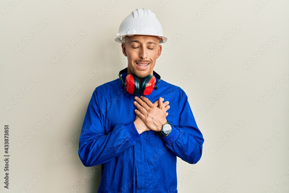 Bald man with beard wearing builder jumpsuit uniform and hardhat smiling with hands on chest with closed eyes and grateful gesture on face. health concept. Bald man with beard wearing builder jumpsuit uniform and hardhat smiling with hands on chest with closed eyes and grateful gesture on face. health concept.
