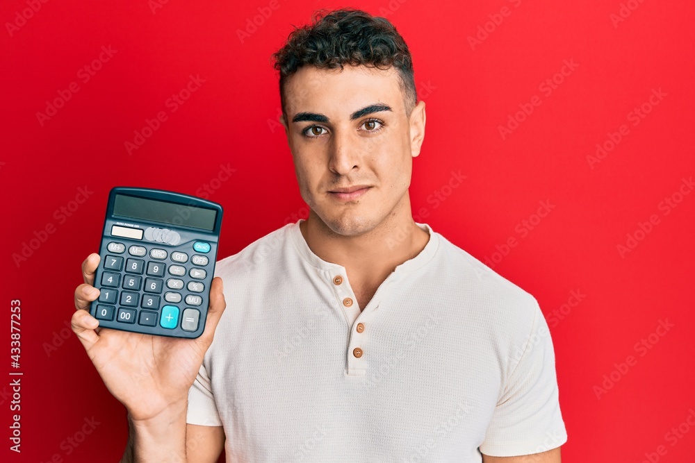 Hispanic young man showing calculator device thinking attitude and sober expression looking self confident Hispanic young man showing calculator device thinking attitude and sober expression looking self confident