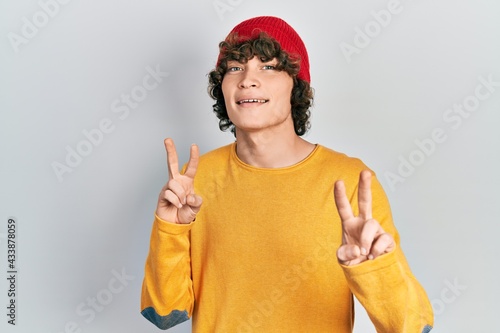 Handsome young man wearing wool hat smiling looking to the camera showing fingers doing victory sign. number two.