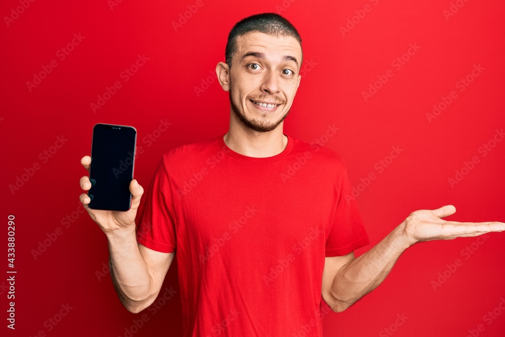 Hispanic young man holding smartphone showing blank screen celebrating achievement with happy smile and winner expression with raised hand Hispanic young man holding smartphone showing blank screen celebrating achievement with happy smile and winner expression with raised hand