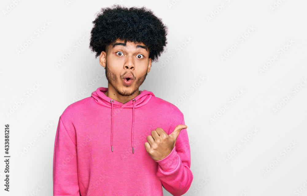Young african american man with afro hair wearing casual pink sweatshirt surprised pointing with hand finger to the side, open mouth amazed expression. Young african american man with afro hair wearing casual pink sweatshirt surprised pointing with hand finger to the side, open mouth amazed expression.