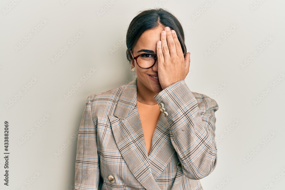 Young brunette woman wearing business jacket and glasses covering one eye with hand, confident smile on face and surprise emotion. Young brunette woman wearing business jacket and glasses covering one eye with hand, confident smile on face and surprise emotion.