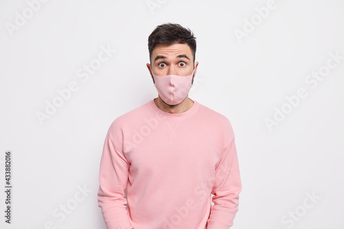 Prevention and safety concept. Surprised young man wears protective mask on face prevents coronavirus spread finds out shocking statistics dressed in pink sweater isolated over white background